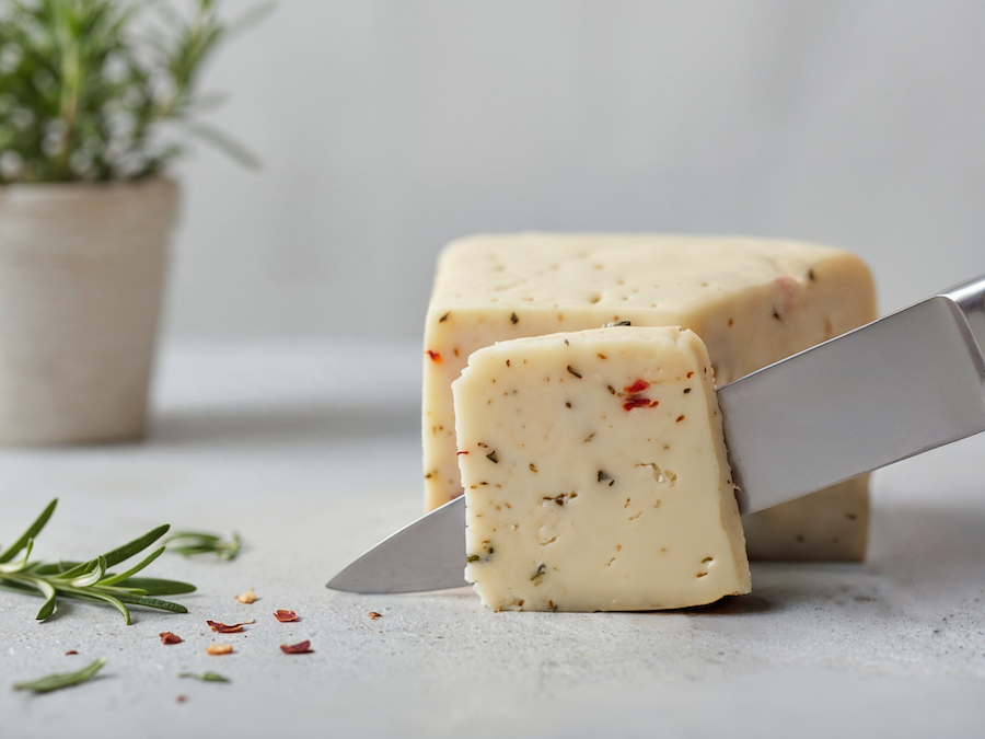 Messer schneidet ein Stück Käse mit Kräutereinlage, daneben Rosmarinzweige