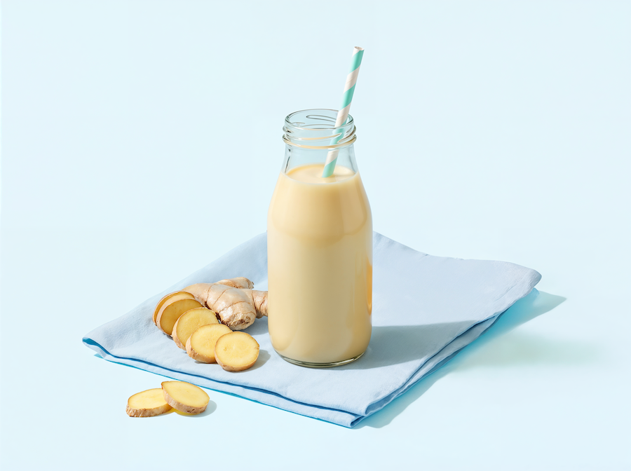 Glasflasche mit cremigem Ingwer-Smoothie und Strohhalm, frische Ingwerscheiben auf blauer Serviette