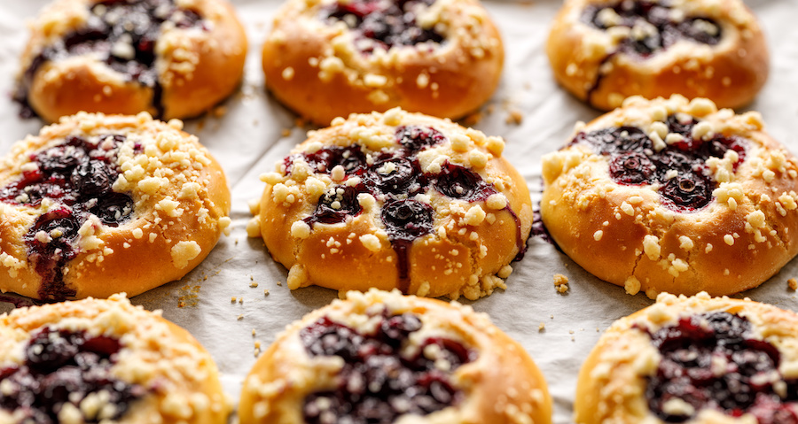 Rows of sweet buns filled with dark berries and topped with crumble on baking paper