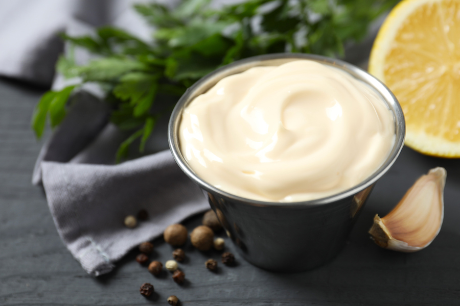 Metallschüssel mit Mayonnaise, daneben Knoblauchzehen, Pfefferkörner, Zitrone und Petersilie auf dunklem Tisch