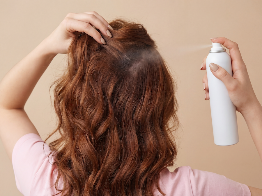Frau mit gewelltem rotem Haar sprüht Trockenshampoo aus einer weißen Dose auf ihren Scheitel