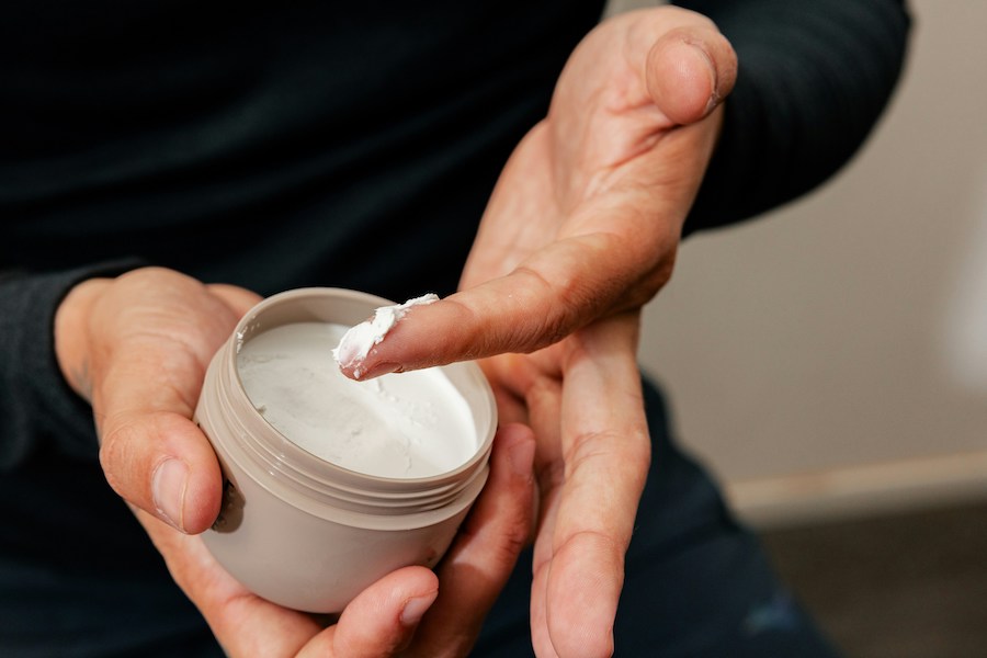 Hände halten einen offenen Tiegel mit weißer Gelcreme und nehmen etwas mit der Fingerspitze