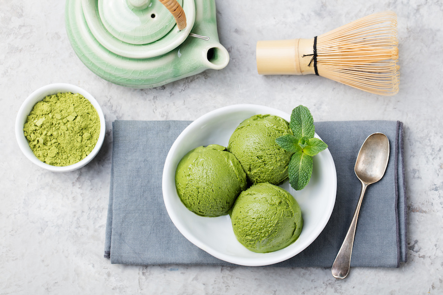Three scoops of matcha ice cream with mint next to a bowl of matcha powder, a teapot and a whisk