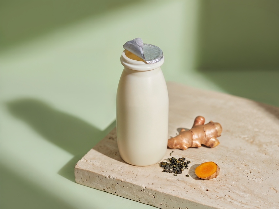 Small white drink bottle with a foil lid next to ginger, turmeric and green tea leaves