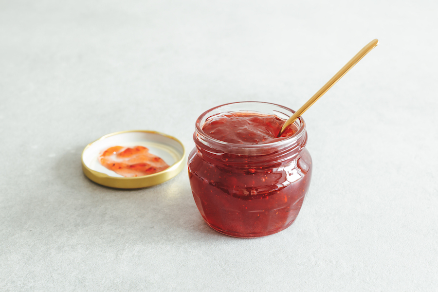 Offenes Glas rote Fruchtmarmelade mit goldenem Löffel neben dem Deckel