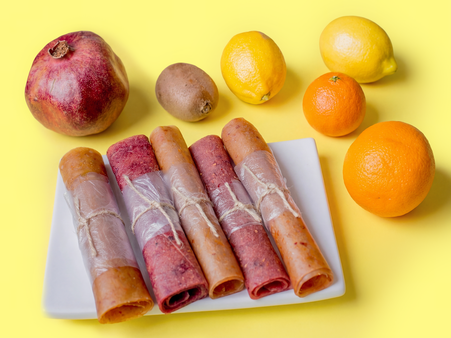 Rolled fruit leather strips on a plate with pomegranate, kiwi, lemons and oranges on a yellow background