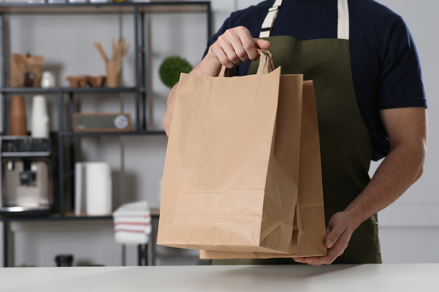 Person in Schürze hält braune Papiertragetaschen an einer Theke