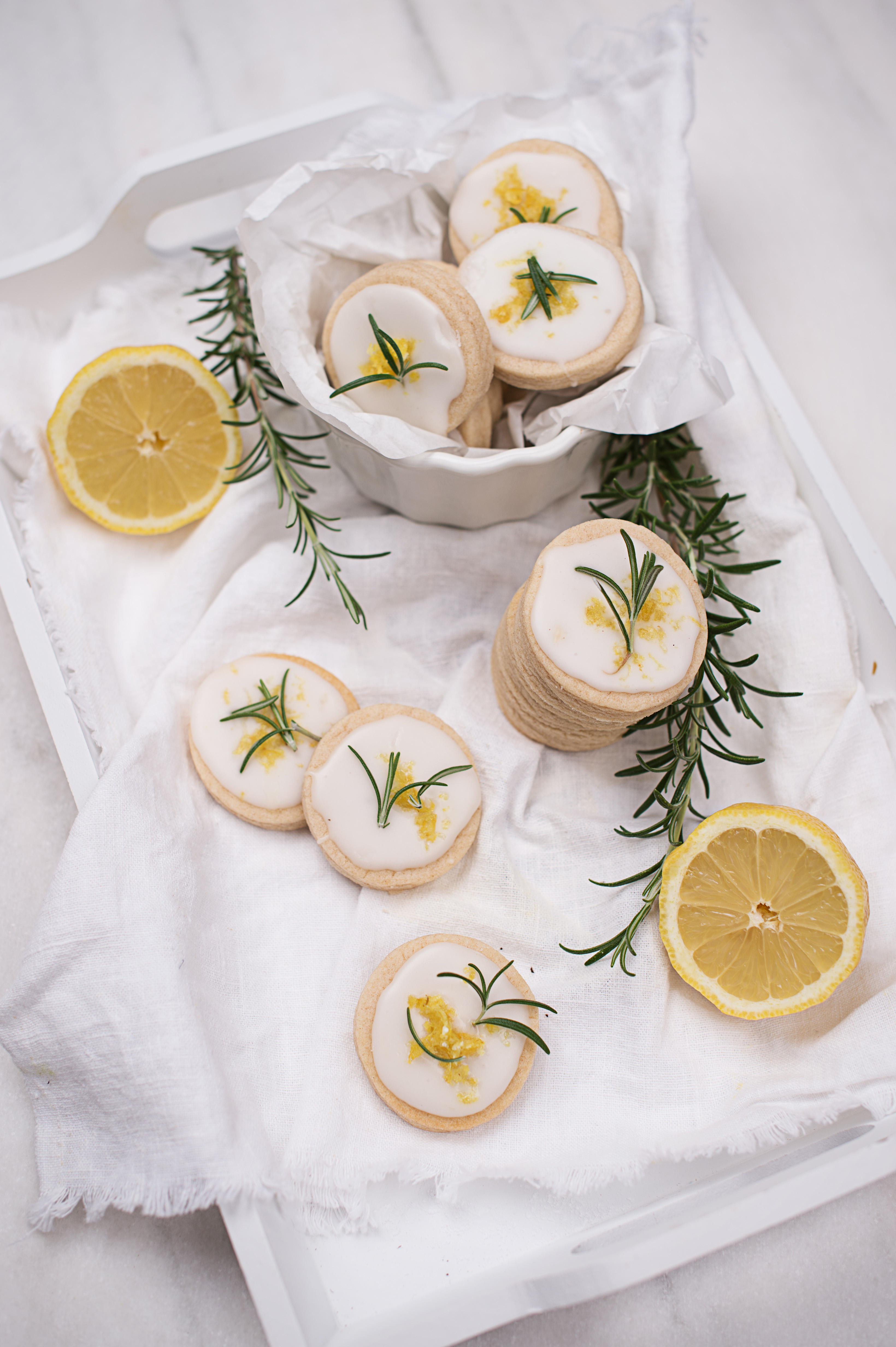 Round shortbread cookies with lemon glaze and rosemary on a white tray with lemon halves [no AI]