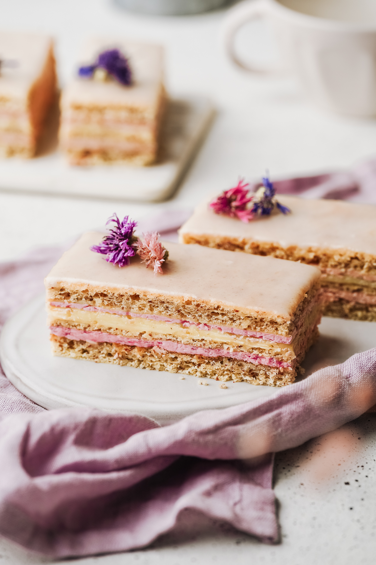 Layered slices with pink cream and vanilla filling under a pale glaze, decorated with edible flowers