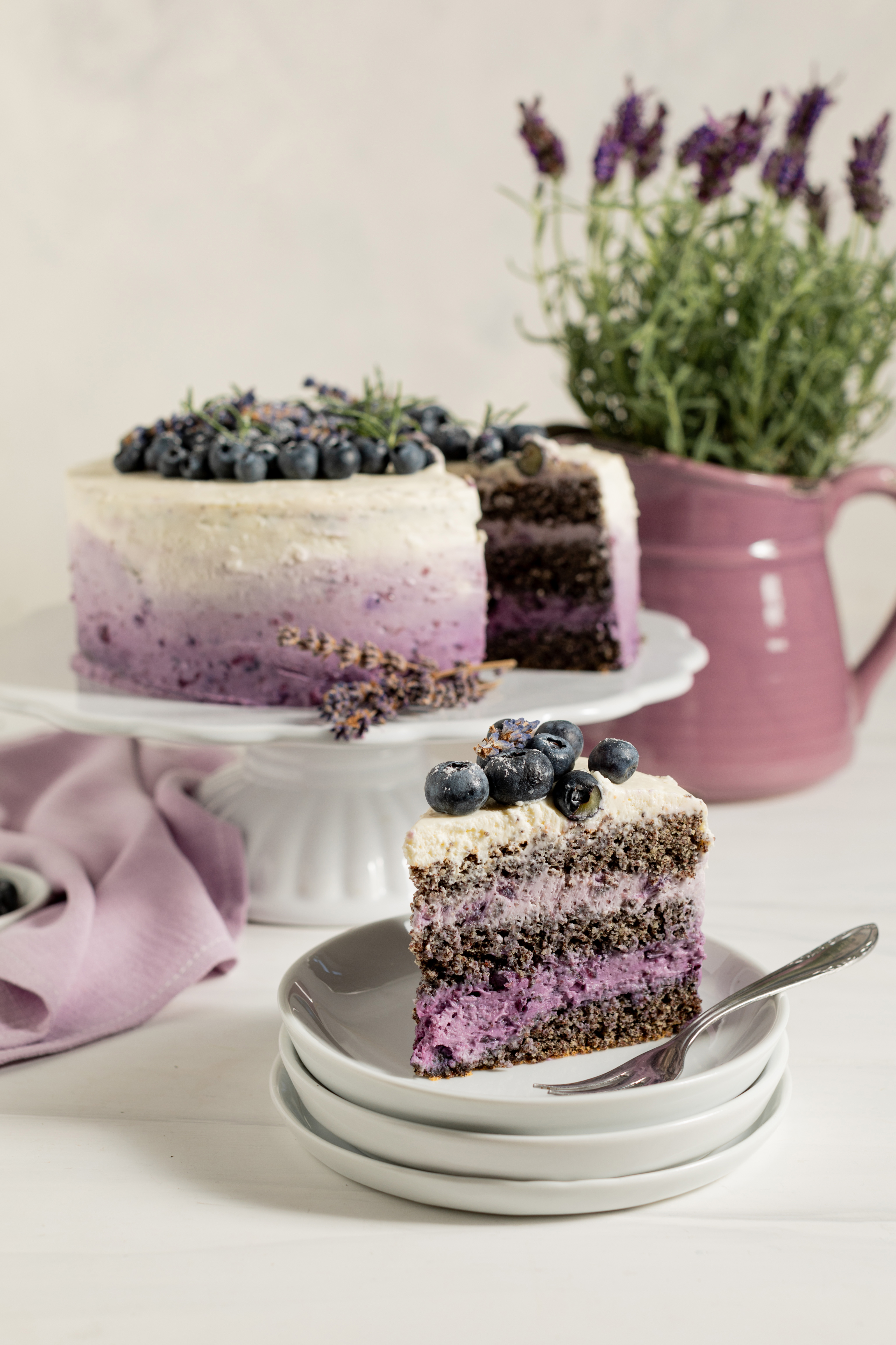 Heidelbeer-Lavendel-Torte mit lila Farbverlauf, ein Stück am Teller und ein Krug mit Lavendel