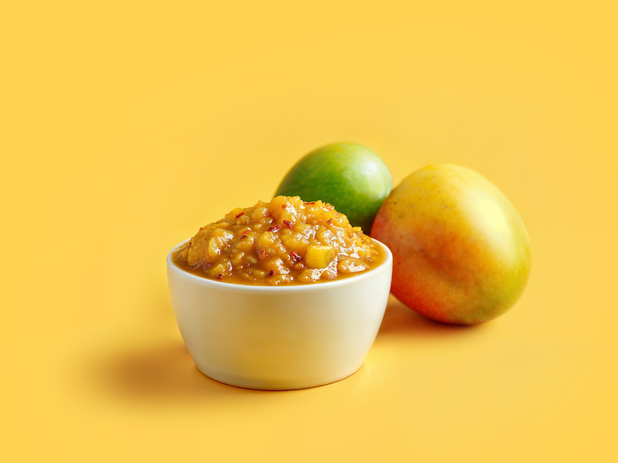 Bowl of chunky mango chutney with two fresh mangoes on a yellow background