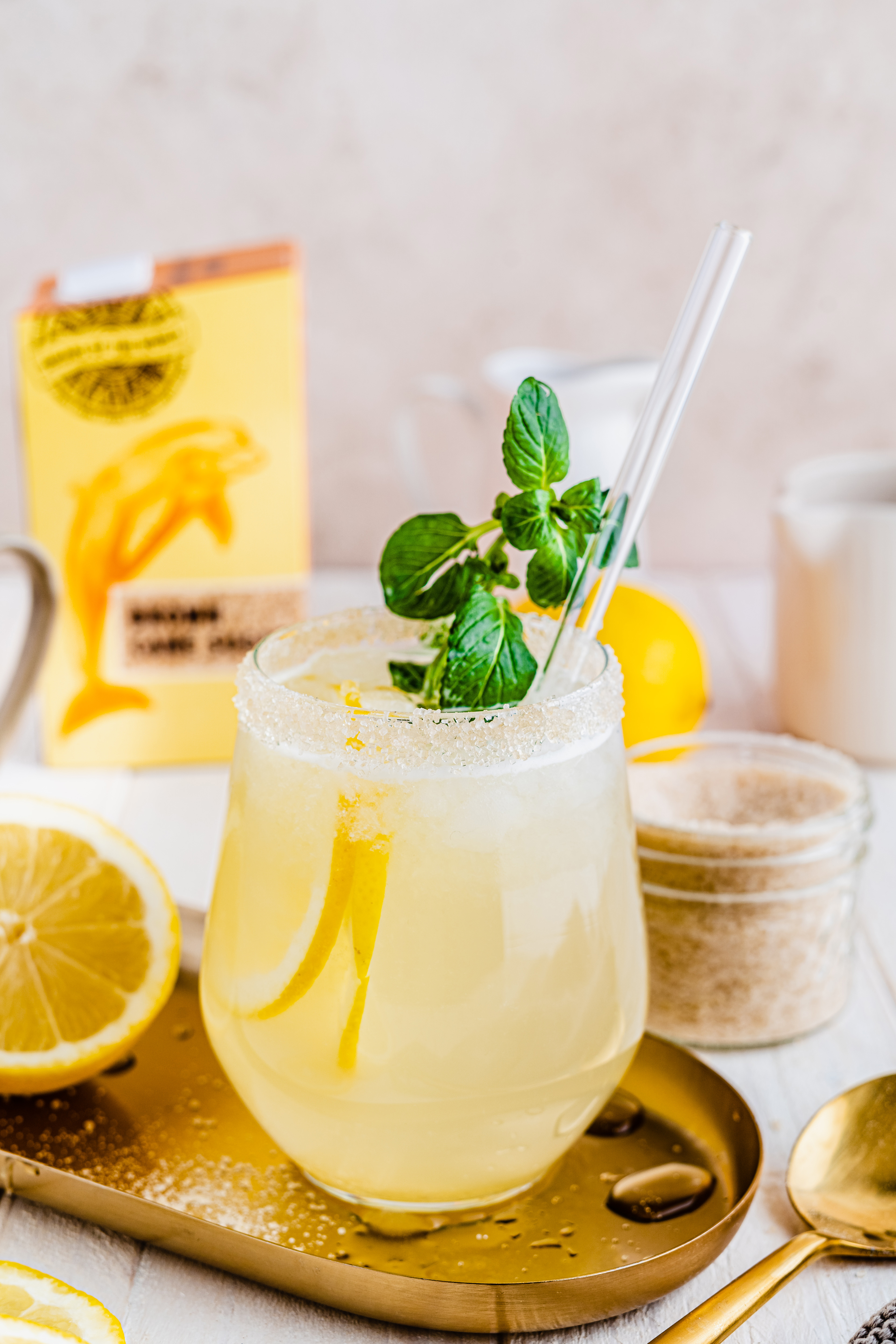 Glas Limonade mit Zuckerrand, Minze und Strohhalm neben einer Zitrone und einem Glas braunem Rohrzucker