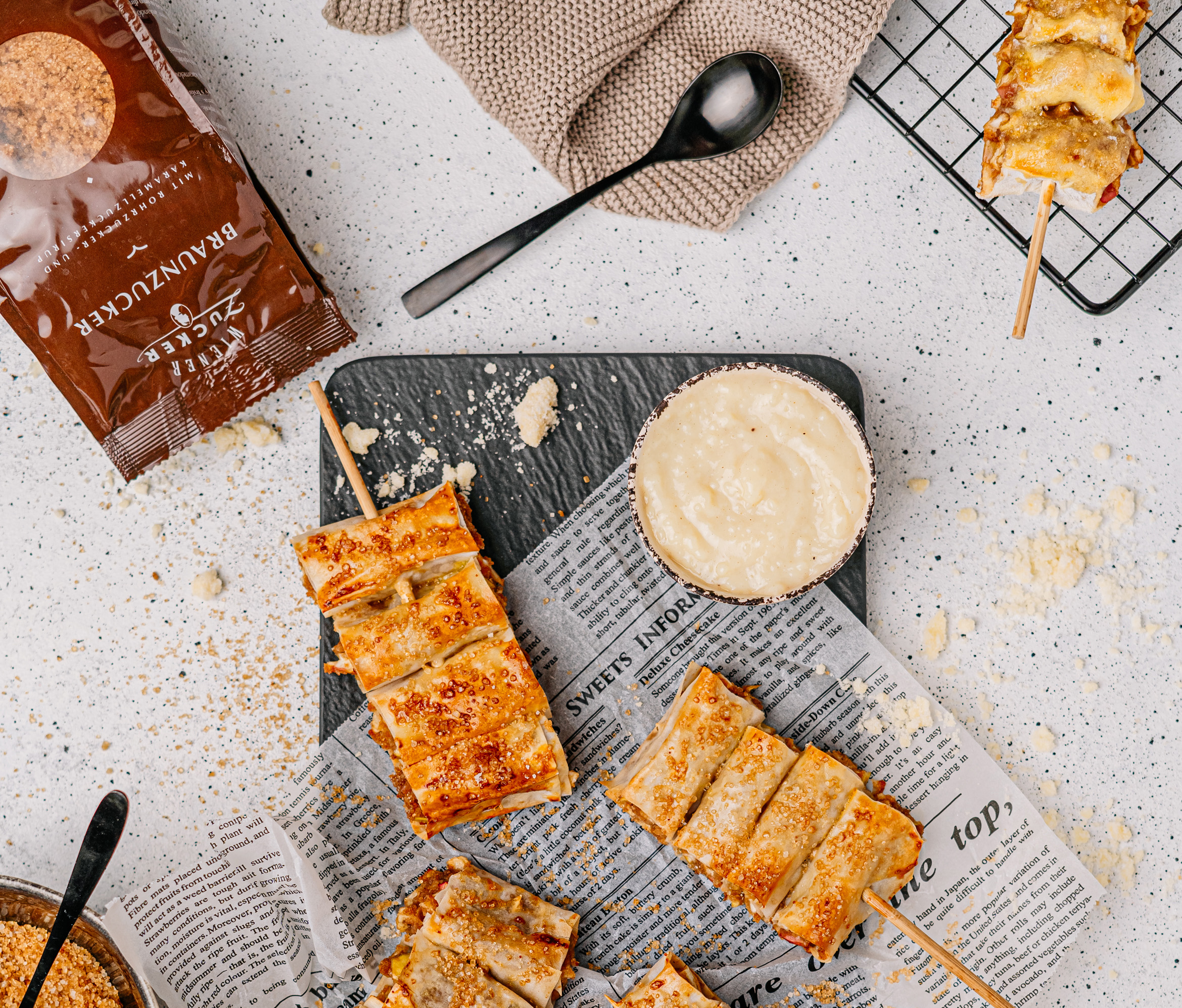 Gebackene Apfelstrudel-Stücke am Spieß mit einer Schale Creme, daneben eine Packung Braunzucker