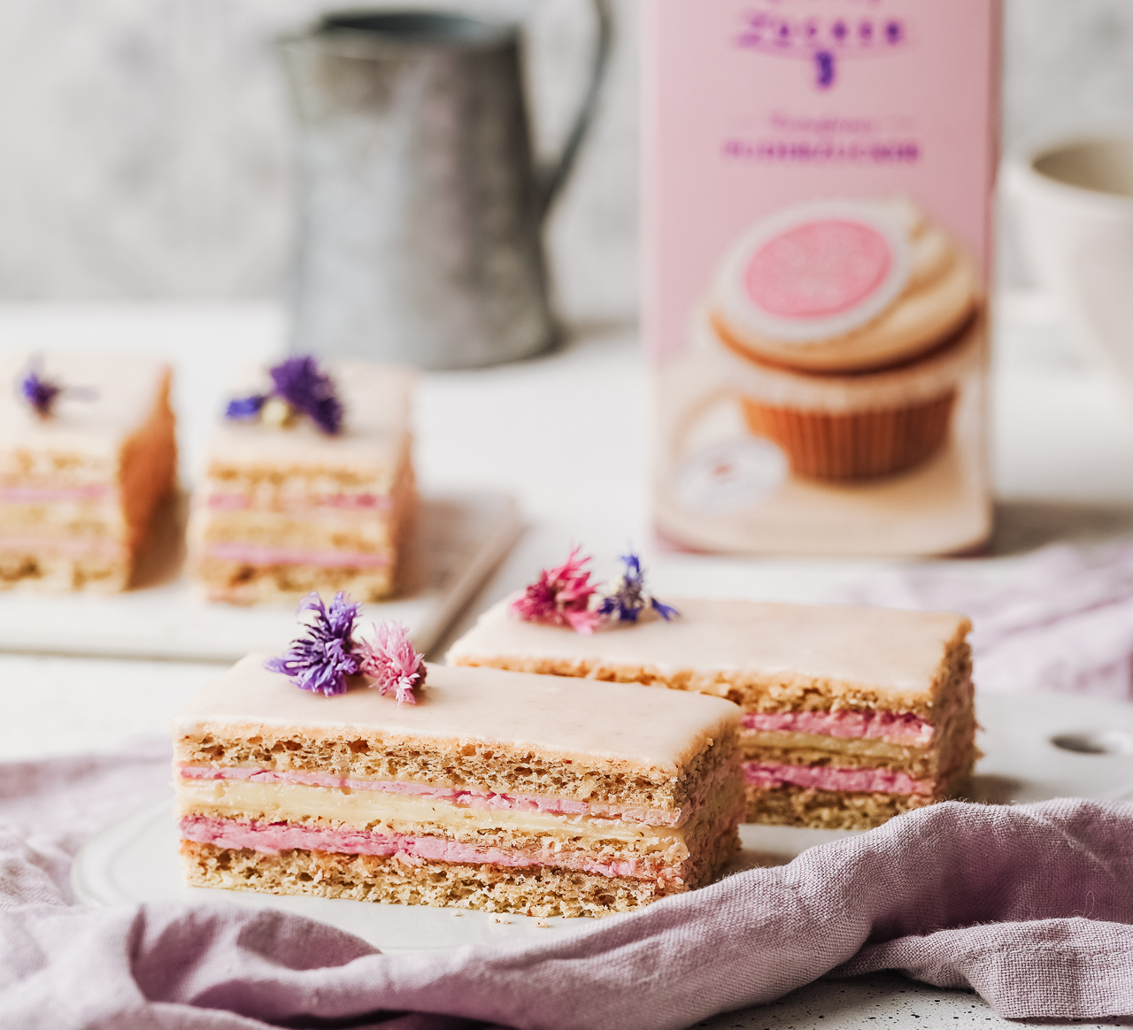 Layered slices with pink cream filling and pale glaze, decorated with edible flowers, sugar pack in the background