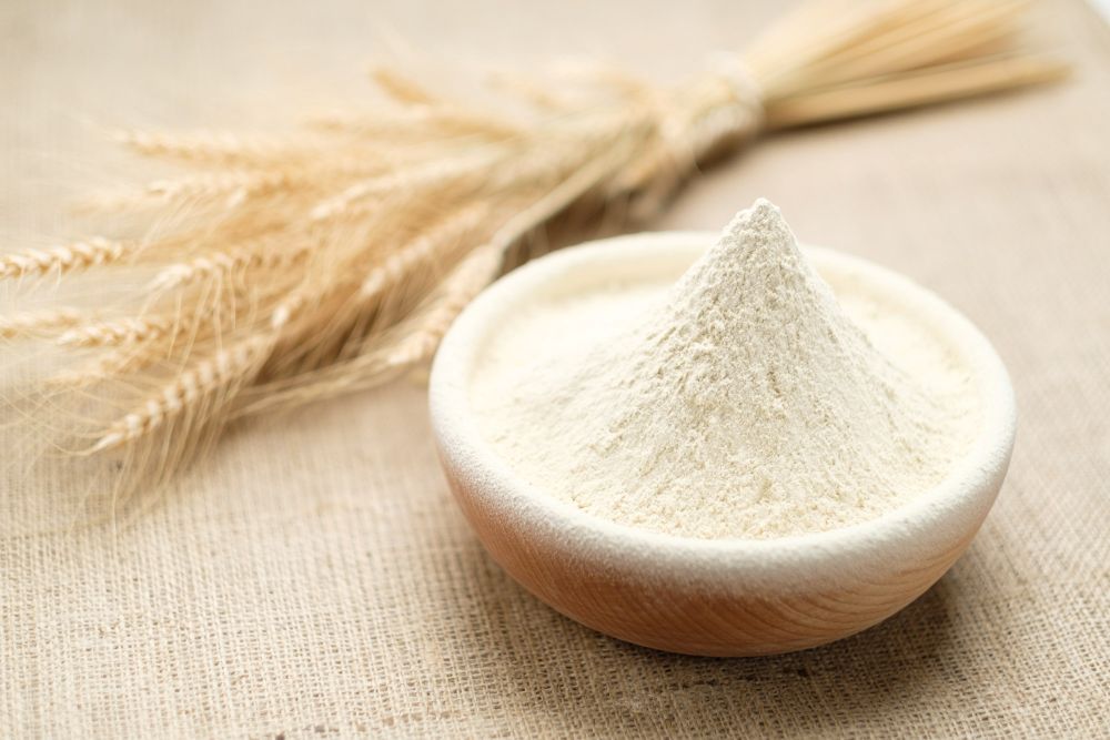 Holzschale mit feinem weißen Weizenpulver neben Weizenähren auf Jute