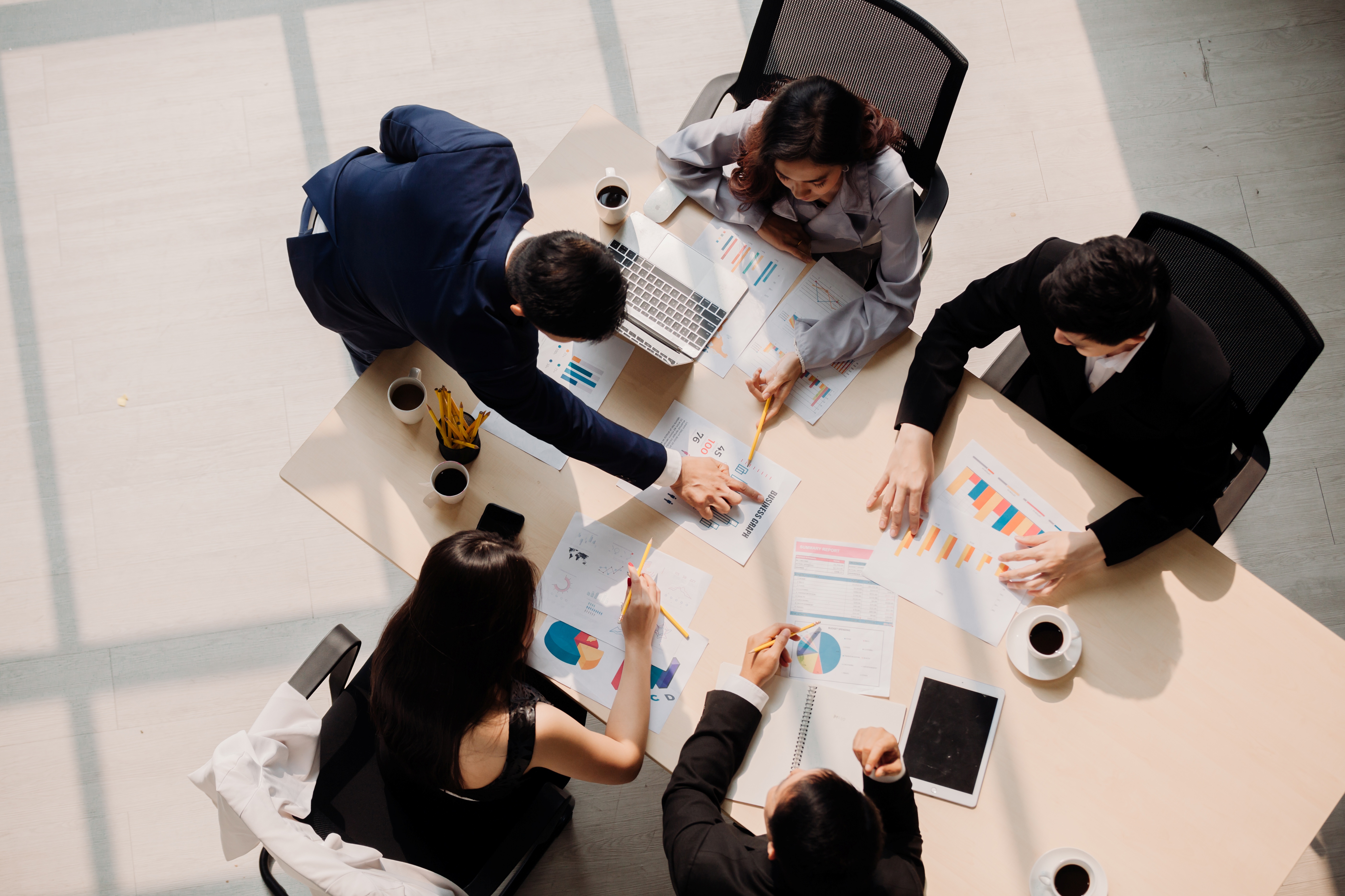 Team of five people working on charts and laptops at a meeting table, seen from above
