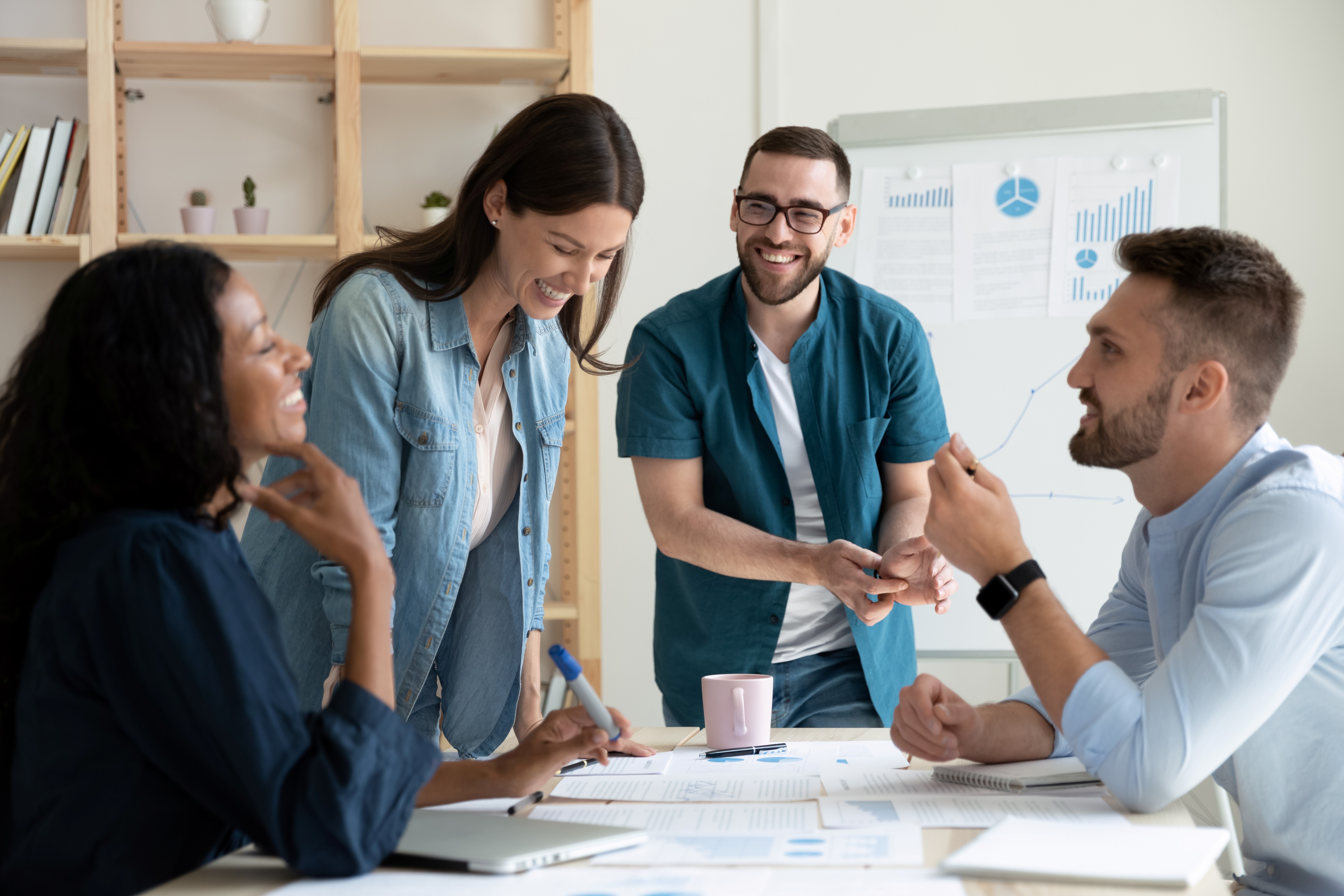 Four colleagues laughing together at a meeting table in front of a whiteboard with charts
