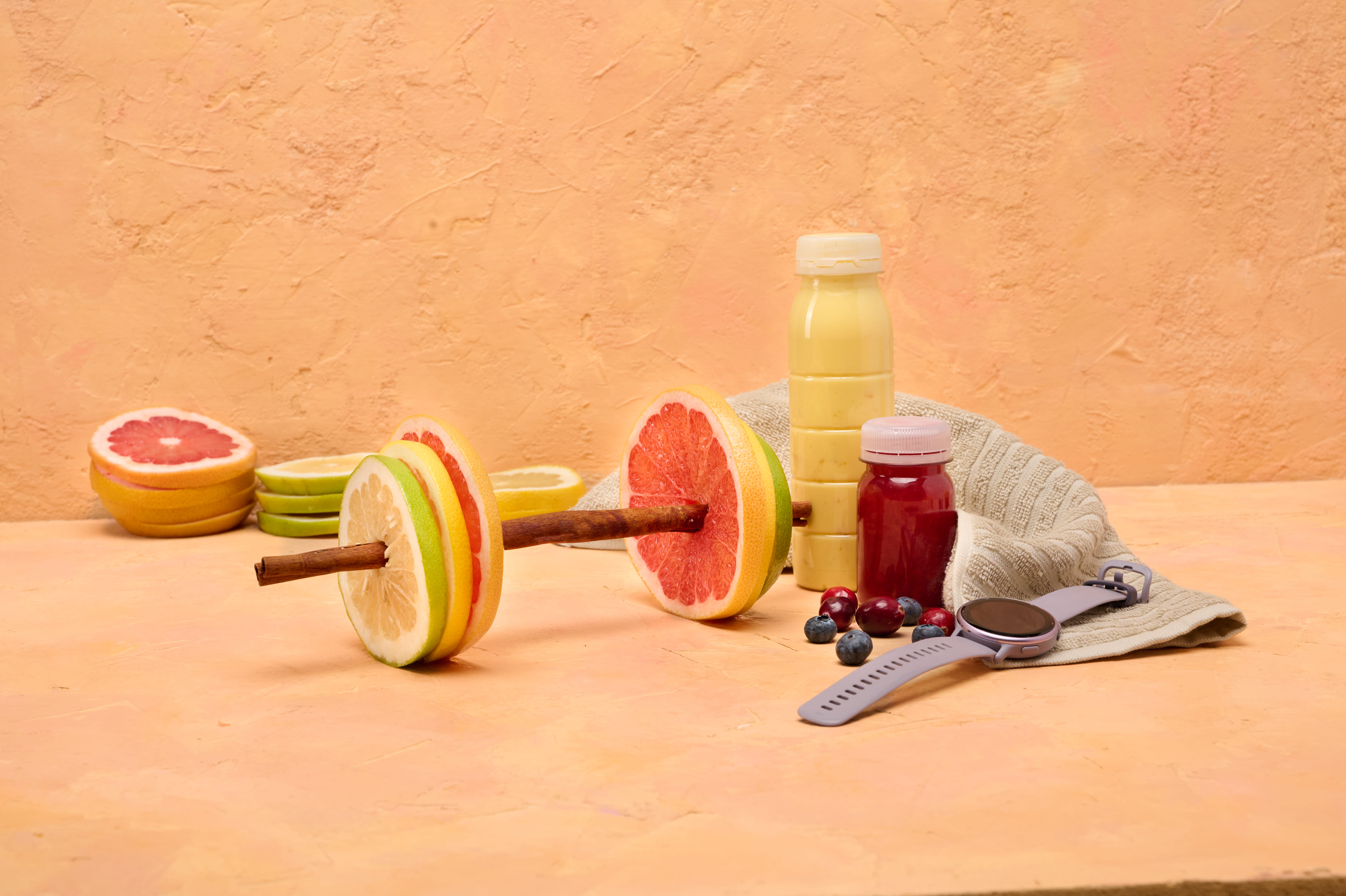 Hantel aus Zitrusscheiben und Zimtstangen neben Saftflaschen, Beeren und einer Fitnessuhr