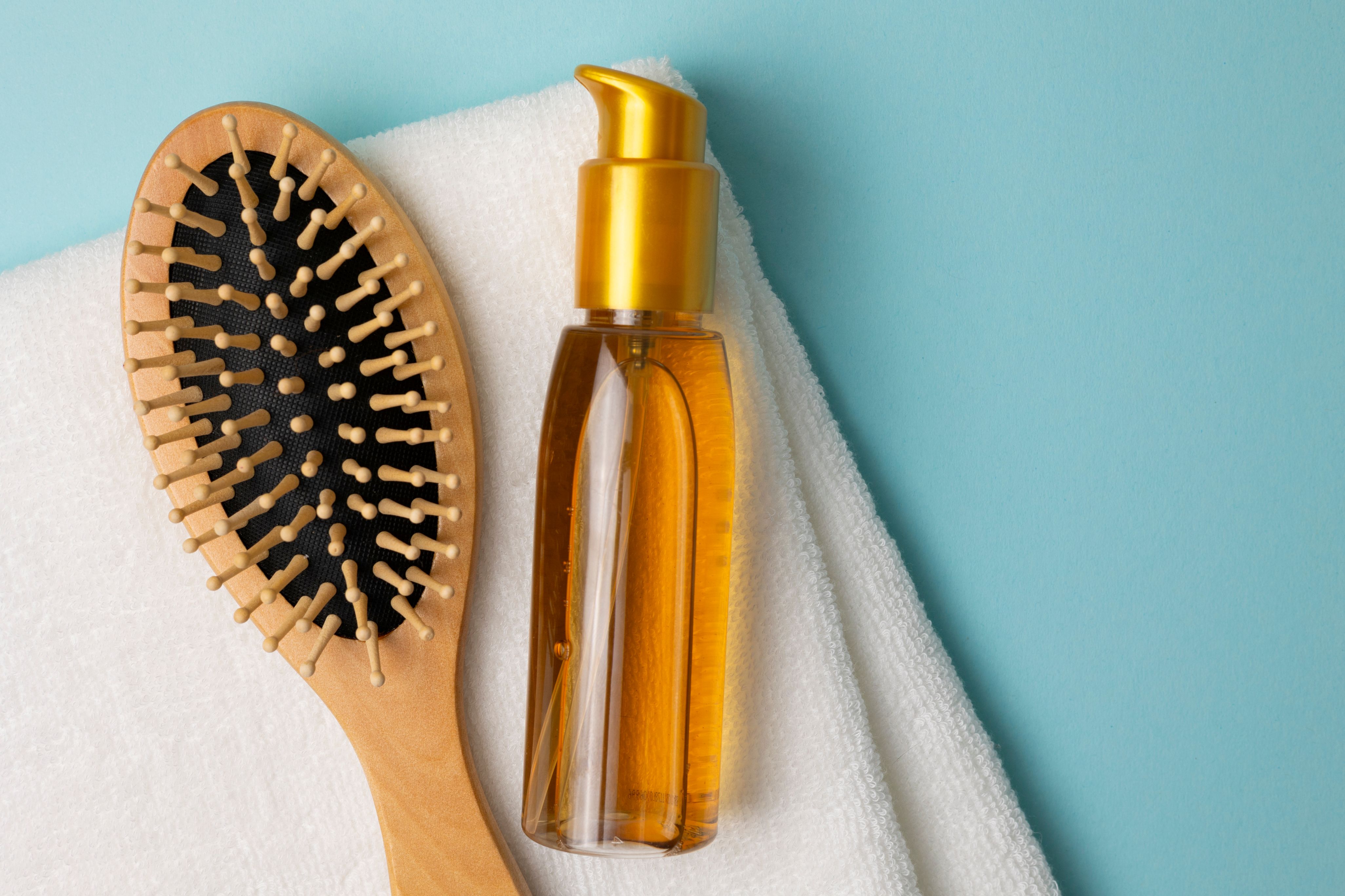 Pumpflasche mit goldenem Haarwasser neben einer Holzhaarbürste auf einem weißen Handtuch