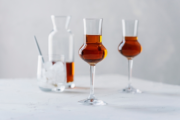 Zwei Stielgläser mit bernsteinfarbener Spirituose vor einer Karaffe und einem Glas mit Eis auf weißem Tisch