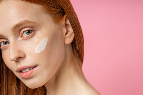 Red-haired woman with a stripe of cream on her cheek against a pink background