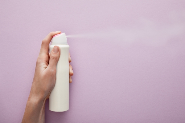 Hand spraying a white aerosol deodorant can against a lilac background