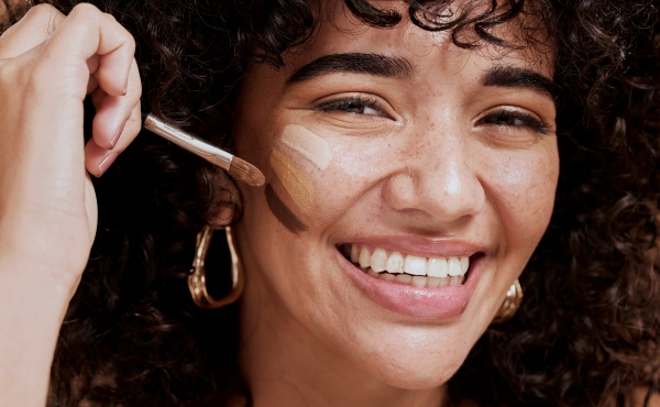 Laughing woman with curly hair applying foundation to her cheek with a brush