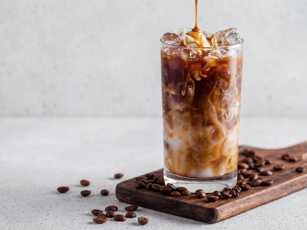 Caramel syrup being poured into a glass of iced coffee with milk, coffee beans on a wooden board