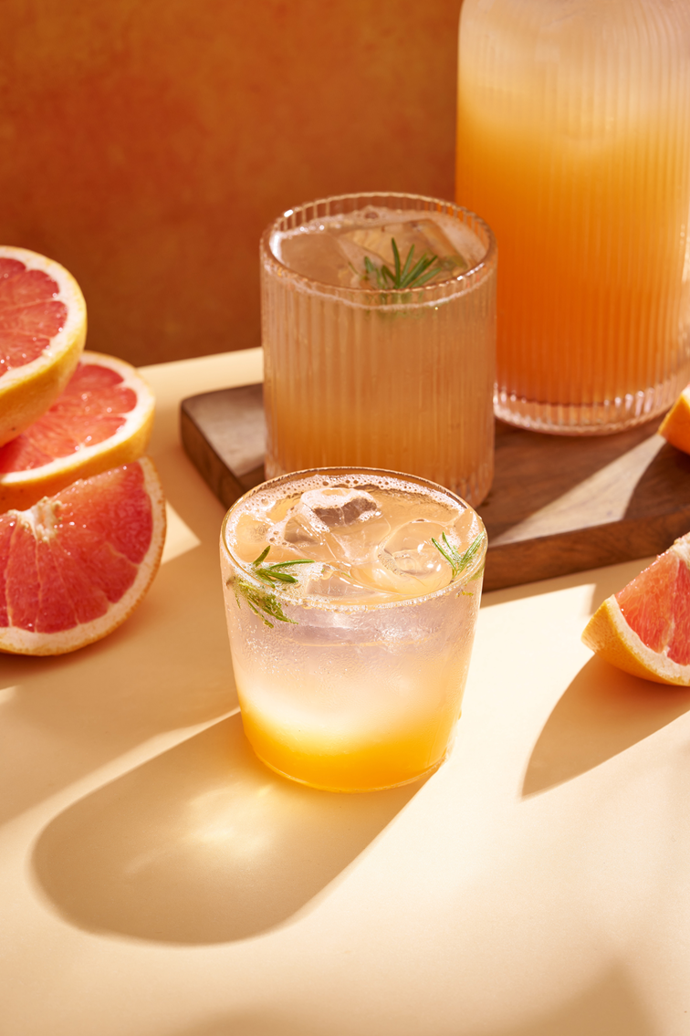 Gläser mit Grapefruit-Drink, Eis und Rosmarin, dazu eine Karaffe und Grapefruithälften im warmen Sonnenlicht