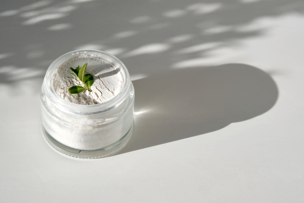 Glastiegel mit weißem Pulverpeeling und grünem Blatt darauf im Schattenspiel der Sonne