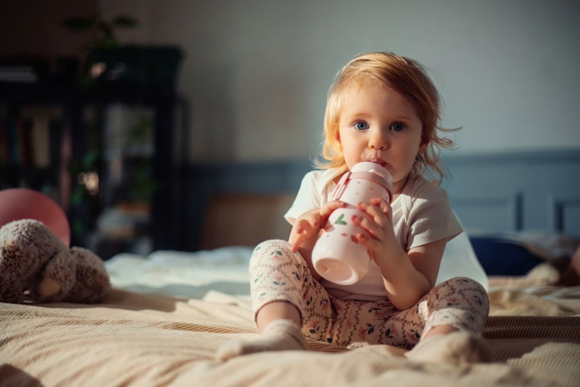 Kleinkind sitzt auf einem Bett und trinkt aus einer Flasche