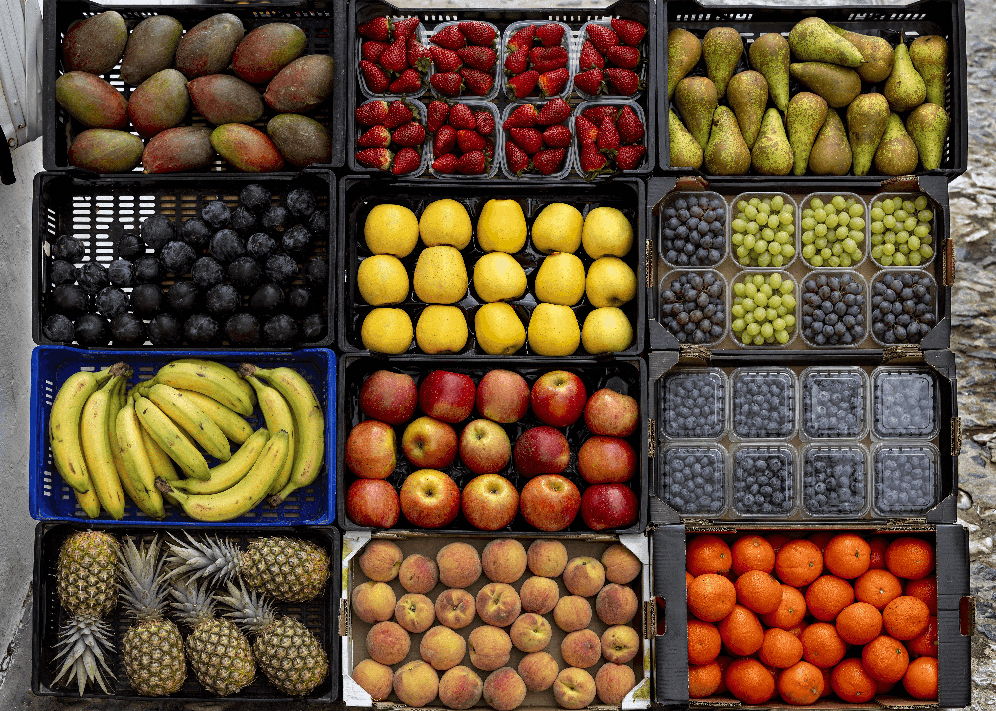 Top-down view of a colorful market-style fruit display arranged in crates. The assortment includes mangoes, strawberries, pears, plums, lemons, red and green grapes, blueberries, bananas, apples, peaches, pineapples, and oranges, neatly organized in rows and containers.