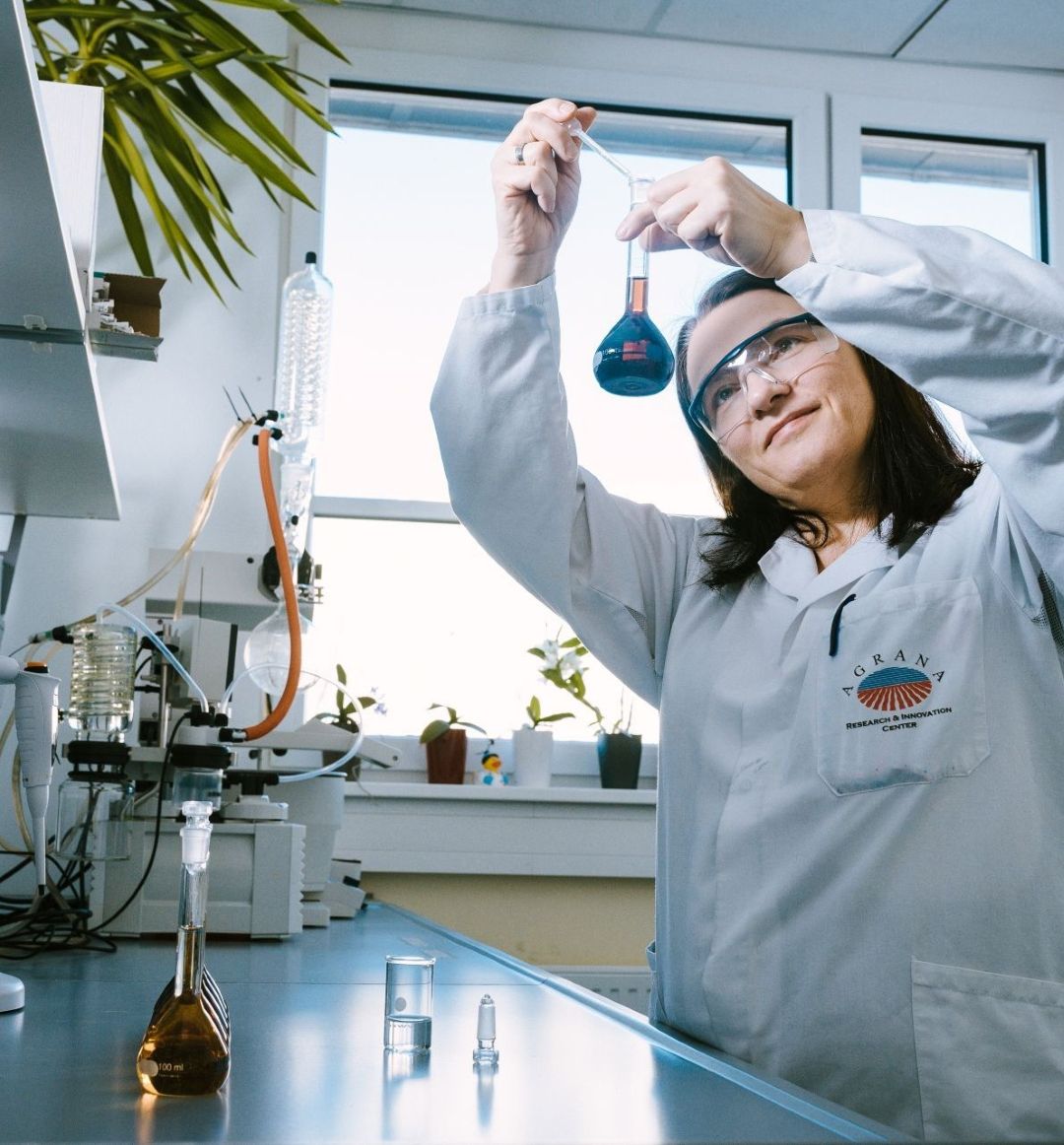 Mitarbeiterin in Laborkittel und Schutzbrille prüft einen Kolben mit blauer Flüssigkeit im AGRANA Research and Innovation Center
