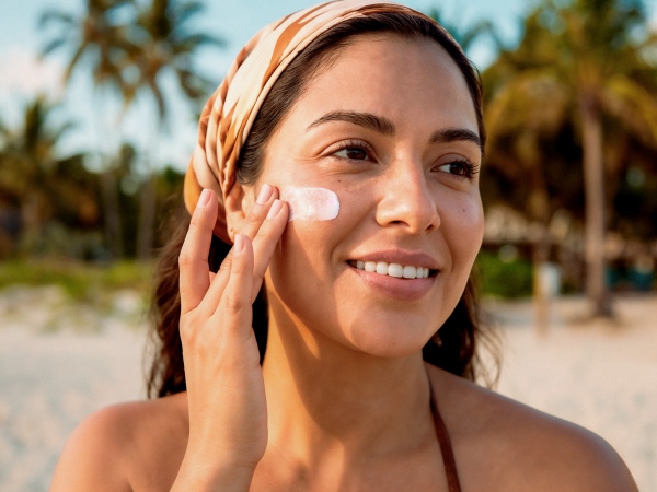 Smiling woman with a headband applying sunscreen to her cheek on a beach with palm trees