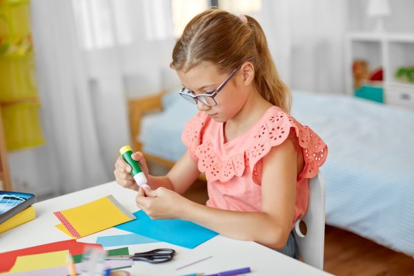 Mädchen mit Brille klebt am Schreibtisch im Kinderzimmer buntes Papier mit einem Klebestift