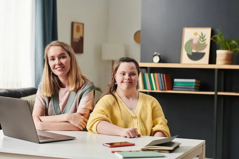Two young women sitting at a desk with a laptop and notebooks.