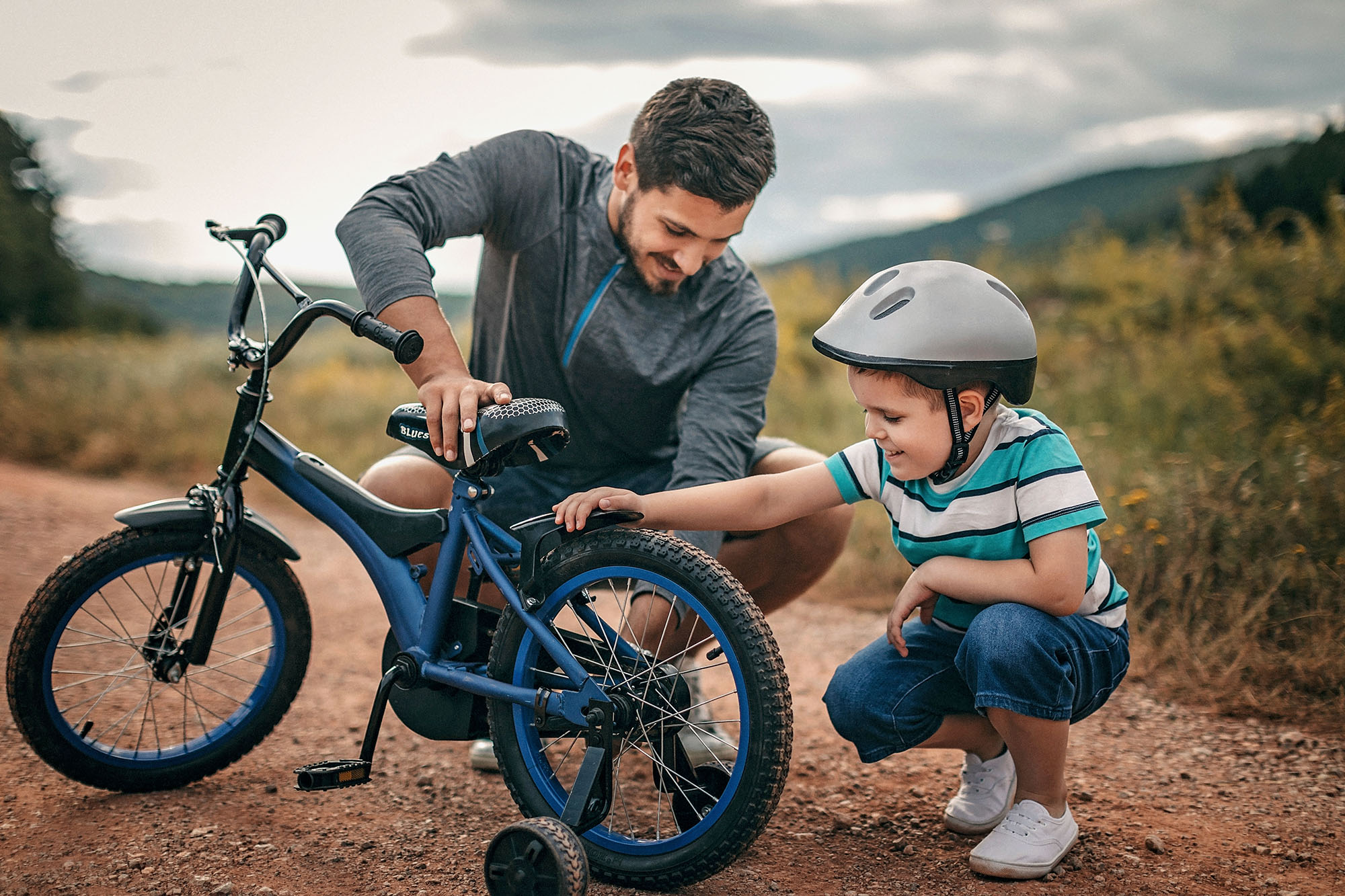 Mann hilft einem kleinen Jungen mit Helm dabei, ein blaues Fahrrad auf einem Feldweg im Freien einzustellen.