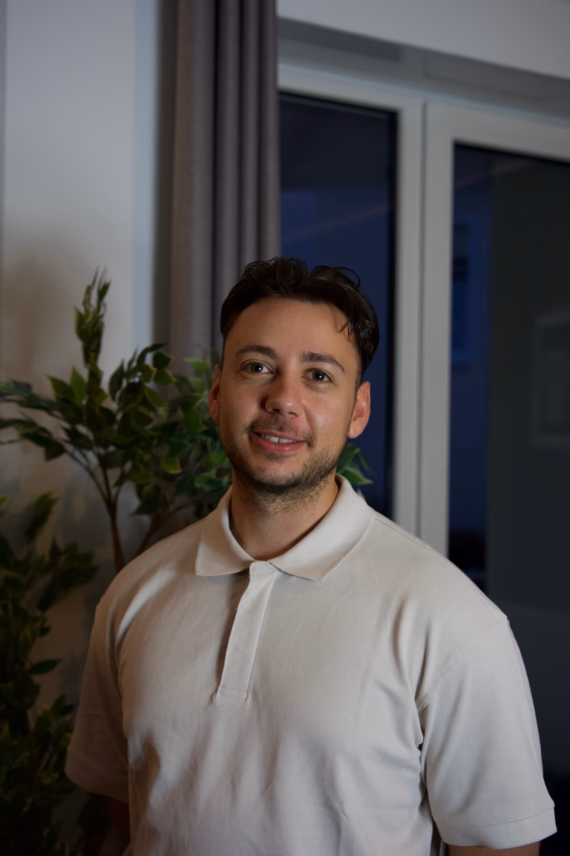 Smiling man wearing a white polo shirt standing indoors near a window at night with green plants in the background.