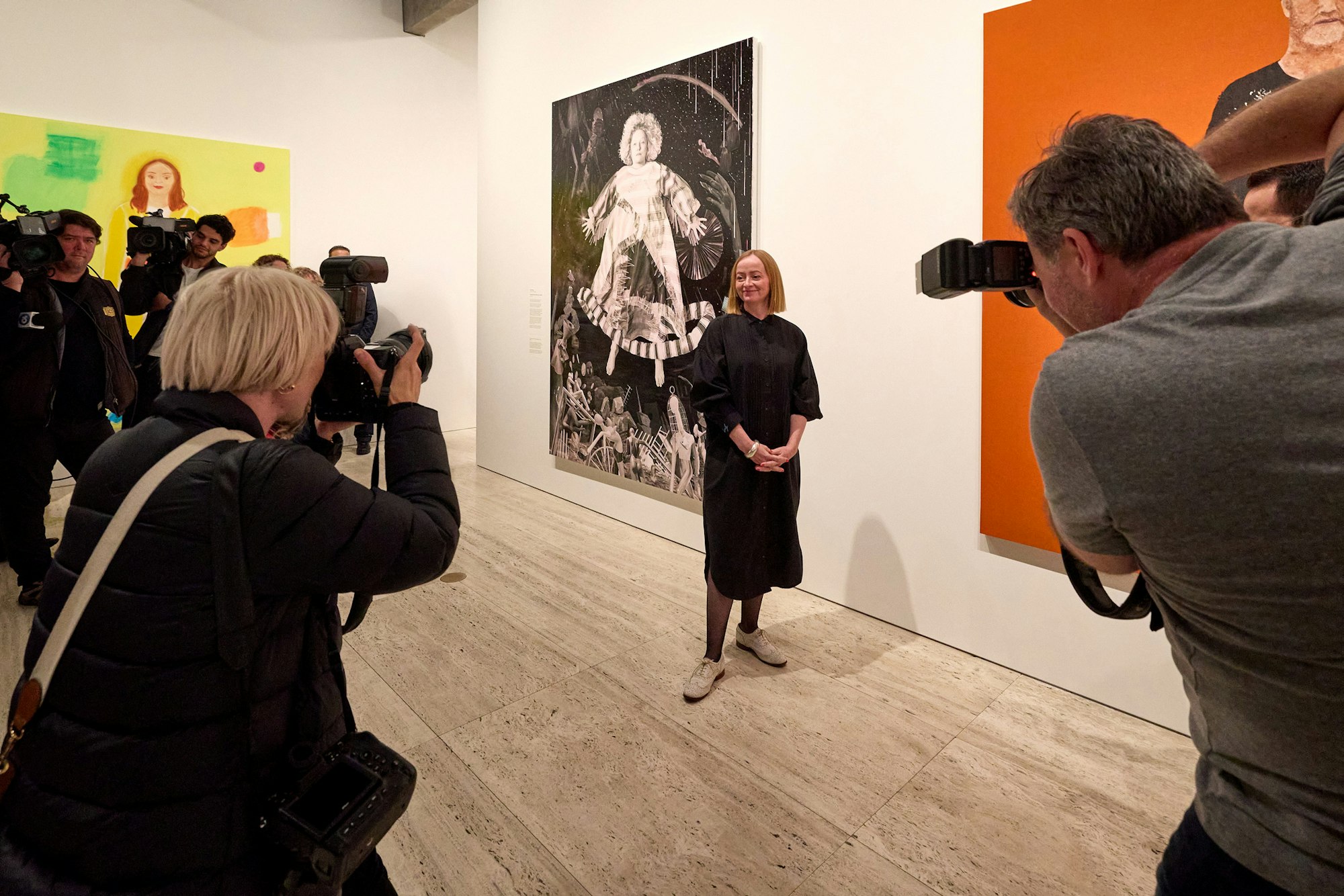 Artist Julie Fragar standing before a large portrait of a woman floating above domestic objects while media photographers capture the scene.