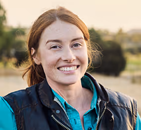 Smiling young woman with red hair wearing a teal shirt and black vest outdoors in natural light.