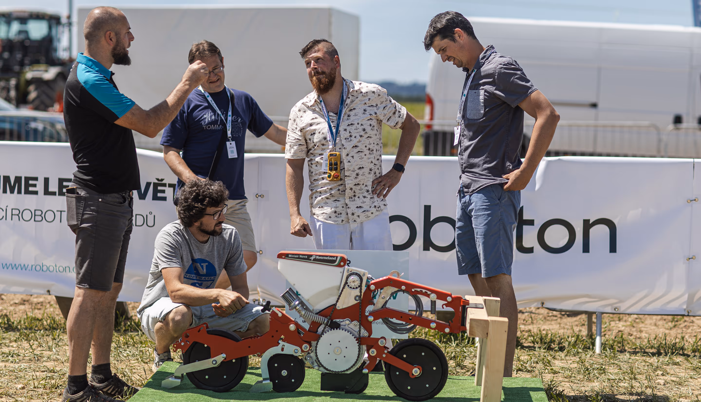 Five men gathered outdoors around a red and white agricultural robot prototype on green turf with a white banner in the background.