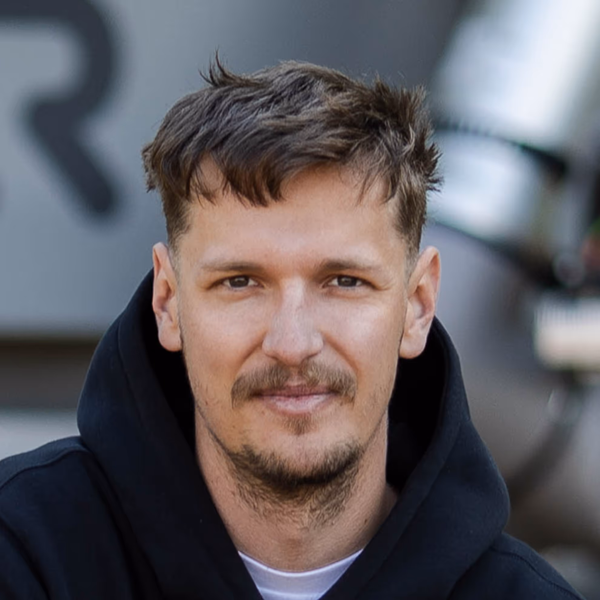 Man with short brown hair and light facial hair wearing a black hoodie, sitting and looking at the camera.