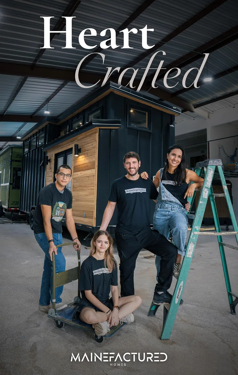 Four young adults posing in front of a tiny house inside a warehouse, two wearing MAINEFACTURED t-shirts, with one sitting on a ladder and another on a dolly.
