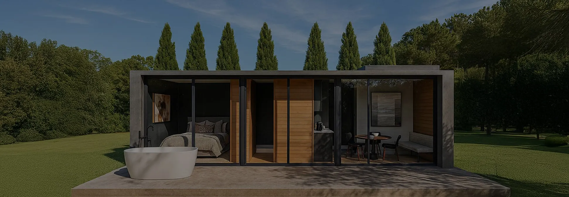 Modern small house with glass walls showing a bedroom, kitchenette, and dining area, set in a green outdoor space with tall trees.