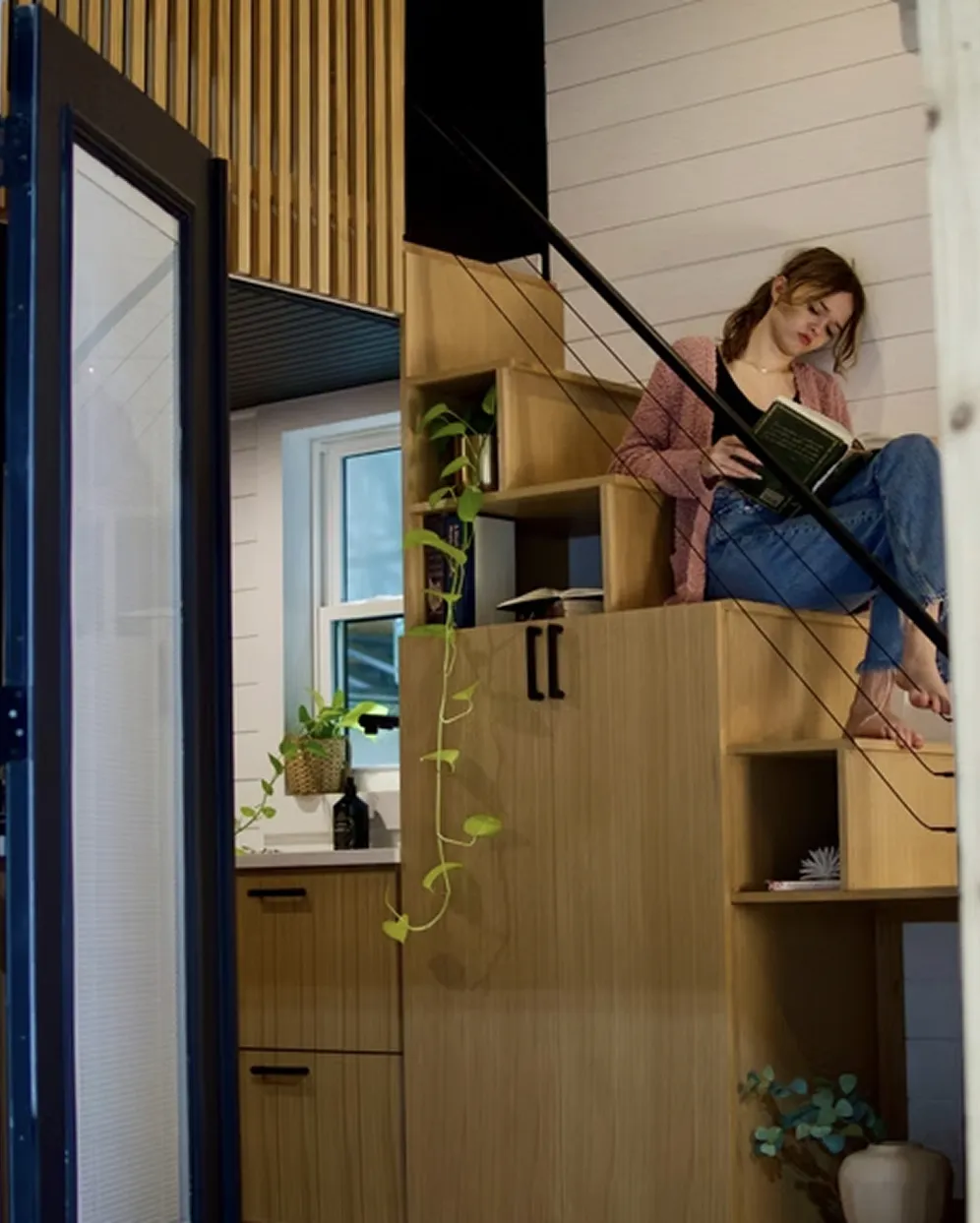 Young woman sitting barefoot on wooden stairs, reading a book indoors by a window with hanging plants nearby.