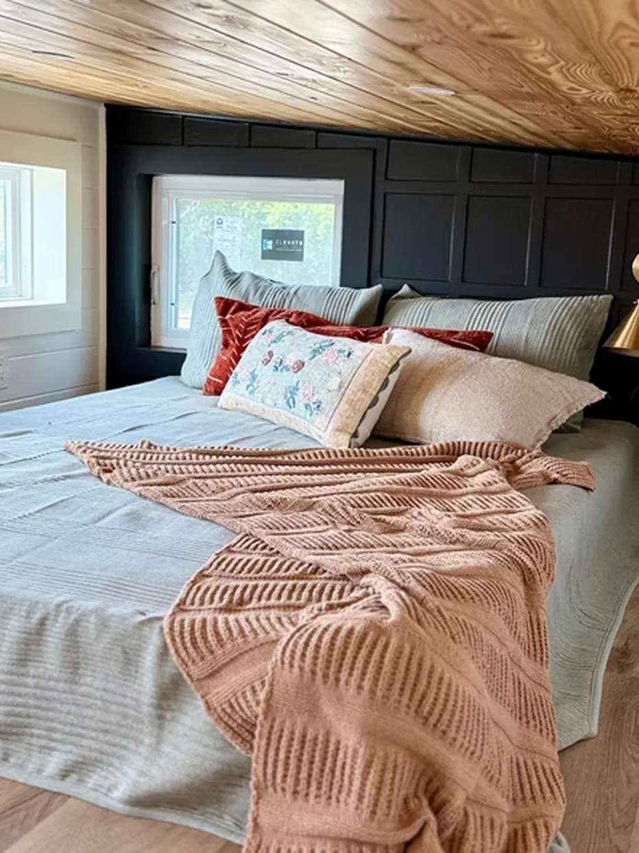 Bed with striped bedding, assorted pillows, and a textured beige throw blanket in a cozy room with a wood-paneled ceiling and window.