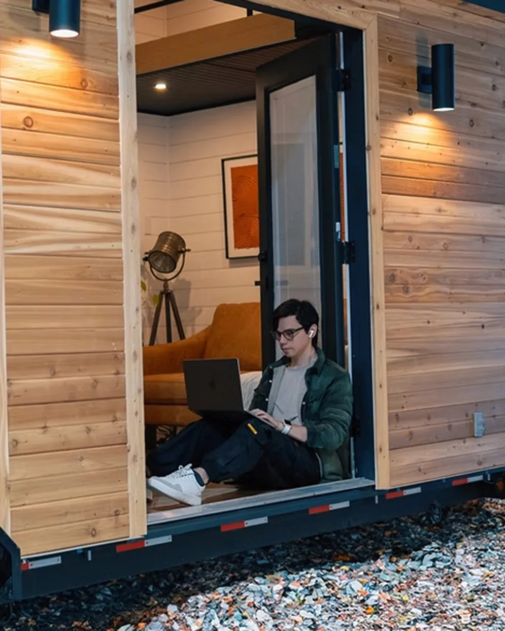 Person sitting at the open doorway of a wooden cabin, working on a laptop with earbuds in.