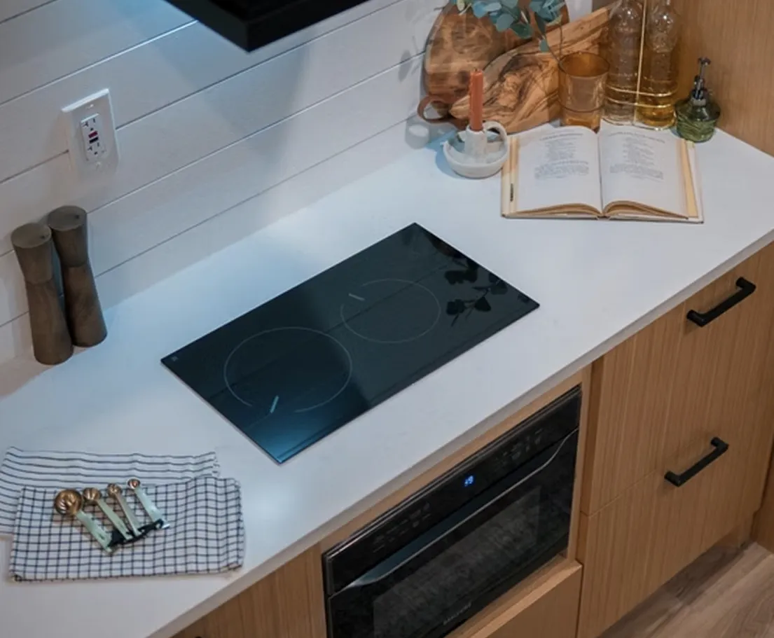 Modern kitchen countertop with a black electric cooktop, wooden salt and pepper mills, an open cookbook, a candle holder, and measuring spoons on a checkered towel.