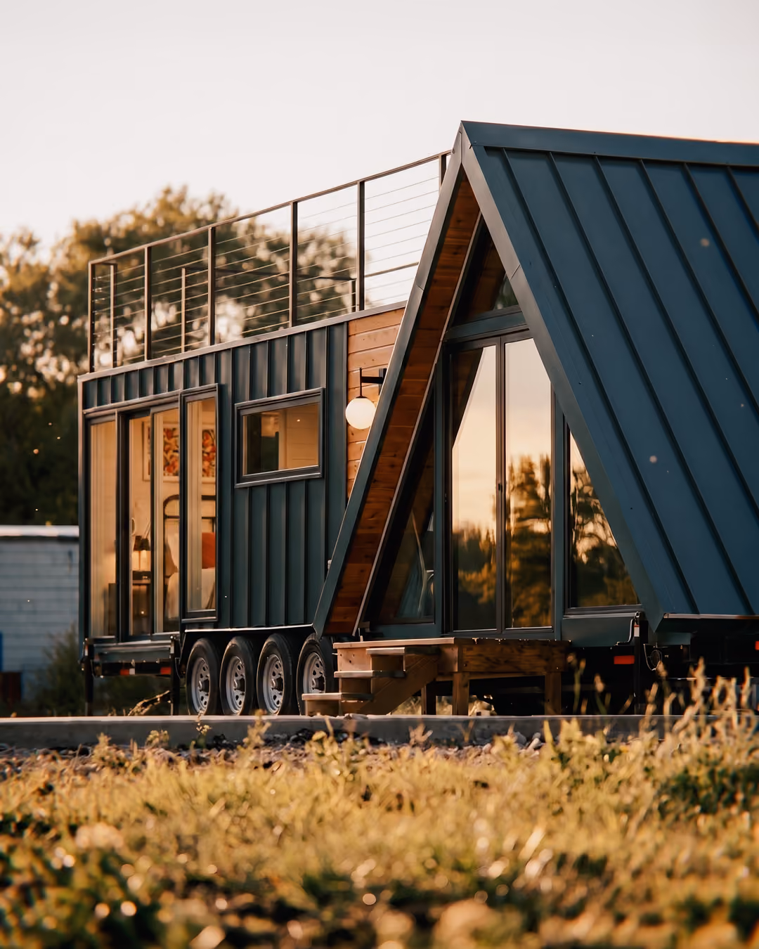 Modern tiny house on wheels with blue metal siding and an A-frame front featuring large glass doors, surrounded by grass and trees at sunset.