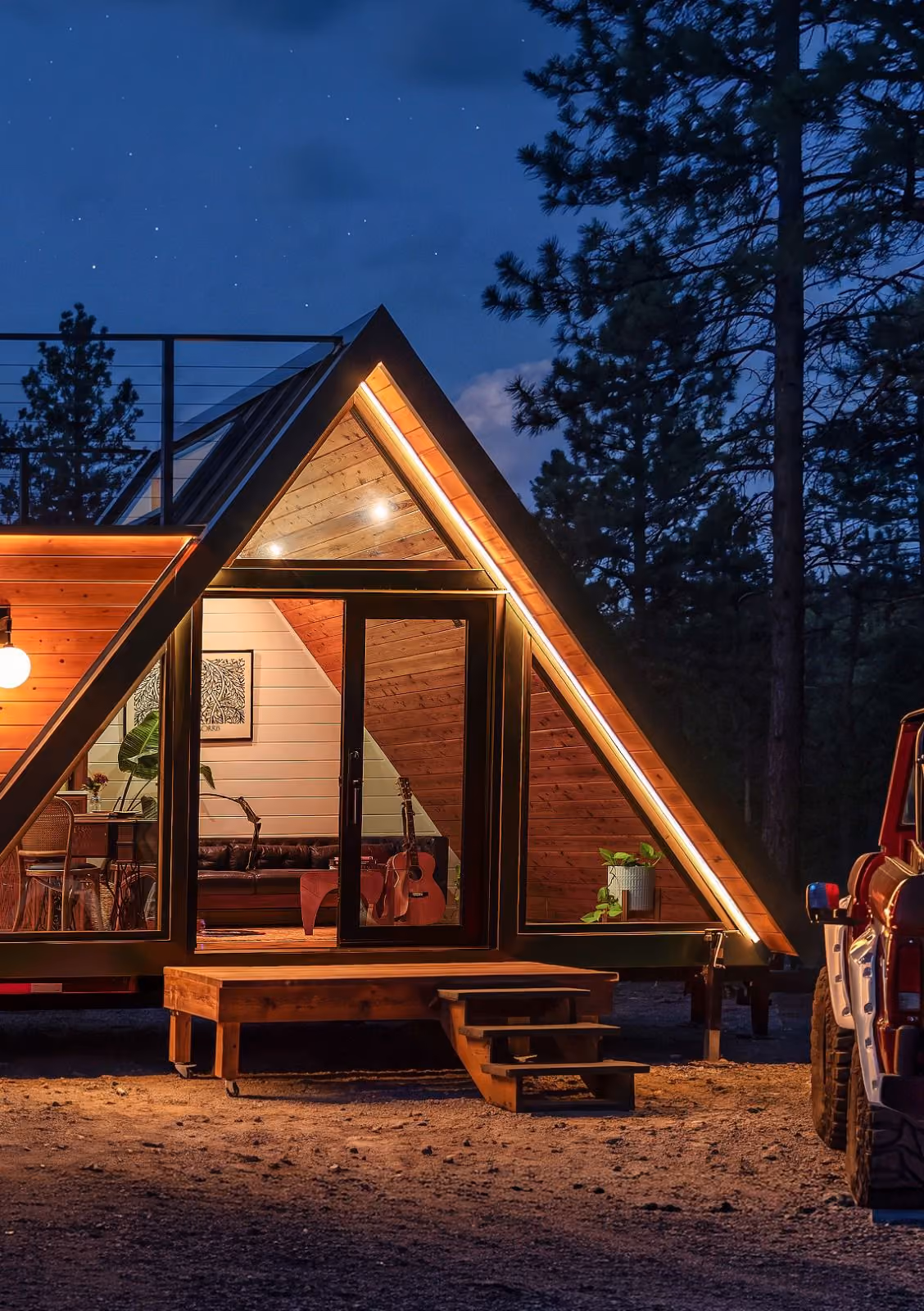 A cozy A-frame cabin illuminated at night with warm lighting, featuring large triangular glass doors, wooden steps leading up to the entrance, a guitar visible inside, and surrounded by tall pine trees under a starry sky.